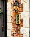 The same three digit address frame is attached to a brick house below a porch light. The frame is made of a light blonde wood and holds peacock blue colored tiles.