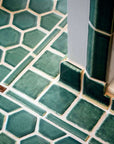 This serene green bathroom floor features hex shaped tiles on the main portion of the floor with square tiles around the edge of the room and a thin border with brown accents that divide the two.