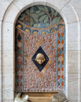 The Pewabic tile drinking fountain sits in a small alcove in a white stone wall. The mosaic tile features circular tiles with harlequin designs on either side. The curved top of the pattern has details of leafy branches with red berries.