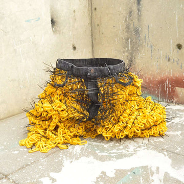 A pair of dark gray jeans rest on the ground. They are sitting upright and open as if someone is sitting in them. The legs have been cut off of the pants and yellow chain-links and pointed black zip ties are attached looking like fungus growing off the the clothing.