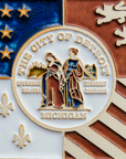 Detail photo of the centerpiece of the Detroit Flag Tile. The circular emblem reads “The City of Detroit” and “Michigan” at the top and bottom. At its center, are two women in red and blue robes. Fire is in the background of the woman on the left and reads “Speramus Meliora” which is latin for “We hope for better things”. The woman on the left is surrounded by a cityscape and reads “Resurget Cineribus” which is “It will rise from the ashes” in latin.