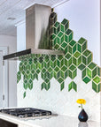 Modern kitchen with range hood against a custom tile backsplash comprised of 4" Rhombus tiles in a gradation of varying green and white glazes. There is a Pewabic Bud Vase with a single yellow flower resting on the glossy white countertop.