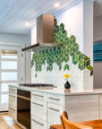 Modern kitchen with range hood against a custom tile backsplash comprised of 4" Rhombus tiles in a gradation of varying green and white glazes. There is a Pewabic Bud Vase with a single yellow flower resting on the glossy white countertop.