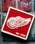 The bright red square Red Wings Tile features the Red Wings logo raised in its center and scraped of glaze giving it a white appearance. There is a simple line border around the tile that is also scraped white. In the bottom right corner are the words "est. 1926".
