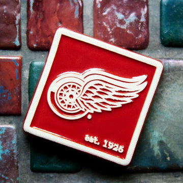The bright red square Red Wings Tile features the Red Wings logo raised in its center and scraped of glaze giving it a white appearance. There is a simple line border around the tile that is also scraped white. In the bottom right corner are the words "est. 1926".