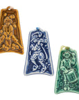 Three colorful embossed ceramic ornaments are on a white background: one shows a man jumping over a stump surrounded by fallen apples, one shows a man dressed in bloomers and a feathered hat as he plays a horn, the last one has a man dressed in Revolutionary War garb as he marches while playing a drum.
