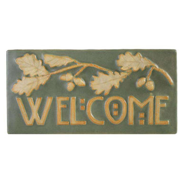 This Oak Welcome Tile features a long oak branch with leaves and acorns. Under the branch is the word "Welcome" in stylized Arts and Crafts style writing. The branch and words are scraped of glaze, giving them a creamy white color while the background is pale green.