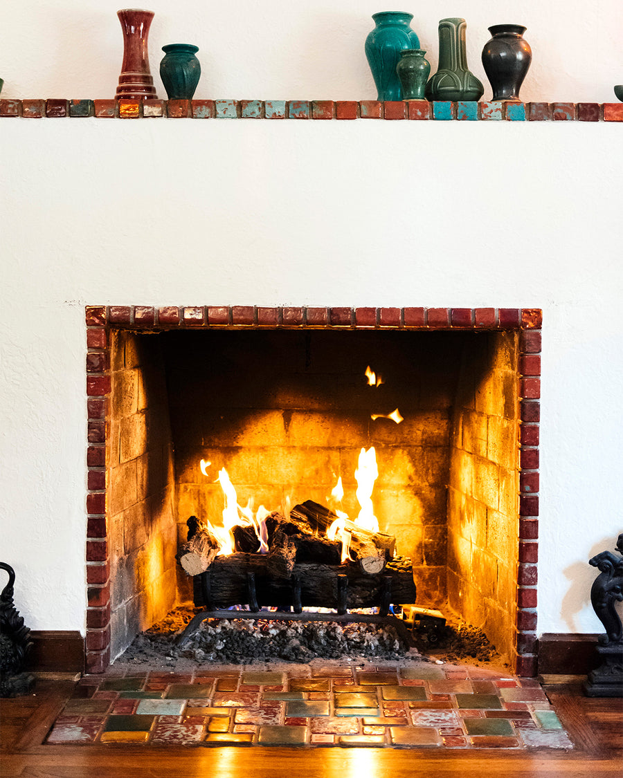 A historic Pewabic fireplace featuring an arrangement of multi-tonal iridescent glazed tiles. The hearth is lit, and there are Pewabic vases on the mantel which also features iridescent tile.