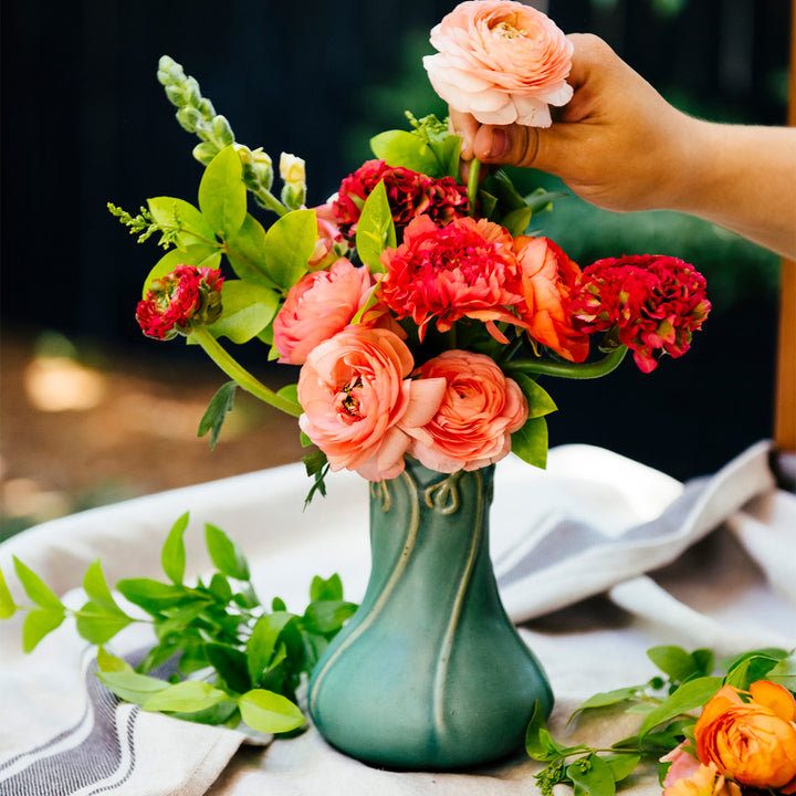 Our Snowdrop Vase in a matte-green "Sorrel" glaze on a light-blue linen fabric. The vase is being filled with bright pink and muted-pink flowers.