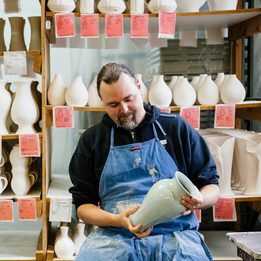 Image of Pewabic's Lead Vessel Maker Andrew holding a Pewabic Classic Vase in a light-blue "Frost" glaze. There are shelves filled with pots waiting to be glazed behind him in our Firehouse work studio.