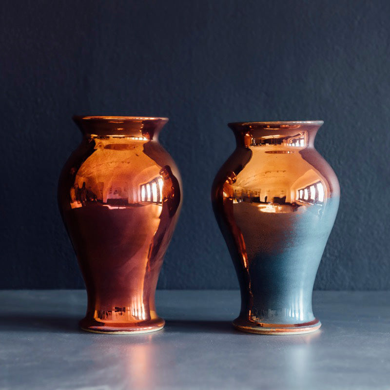 Two copper colored iridescent vases side by side against a dark gray/blue background. The vase on the left looks like a vibrant sunset, while the vase on the right has flashes of muted blue on the surface of a mostly copper base. 