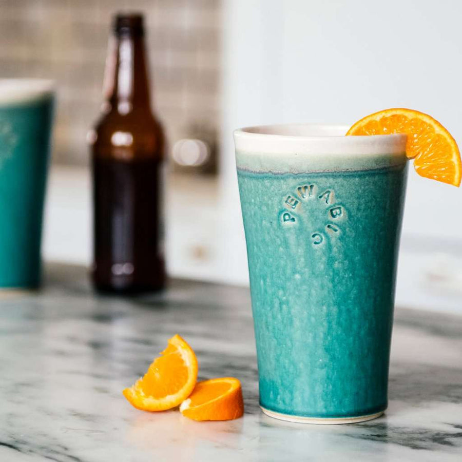 A Pewabic Pint in Pewabic Blue on a classic marble countertop. There is an open bottle of beer and another Pint just out of view. The Pint in-frame has a garnish of fresh orange slices.