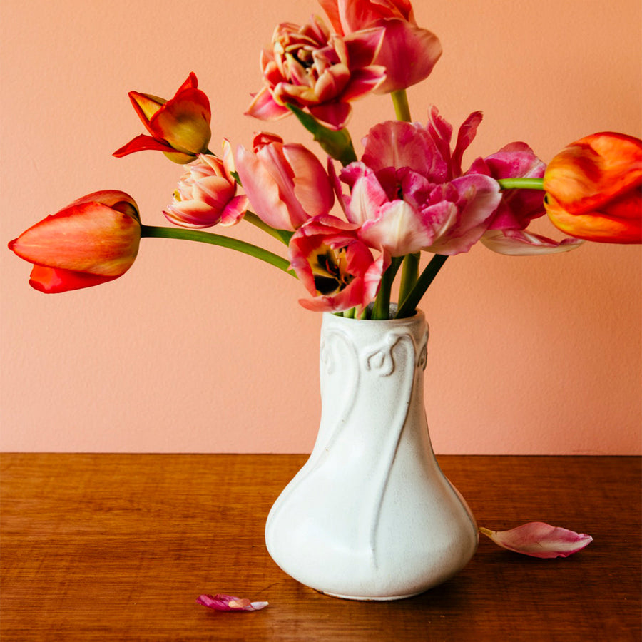  Our Snowdrop Vase in a white, slightly iron-speckled, "Birch" glaze rests on a dark wooden table. The vase is filled with bright-red and pink tulip varieties. The wall behind the vase is a peachy, springy, pink.
