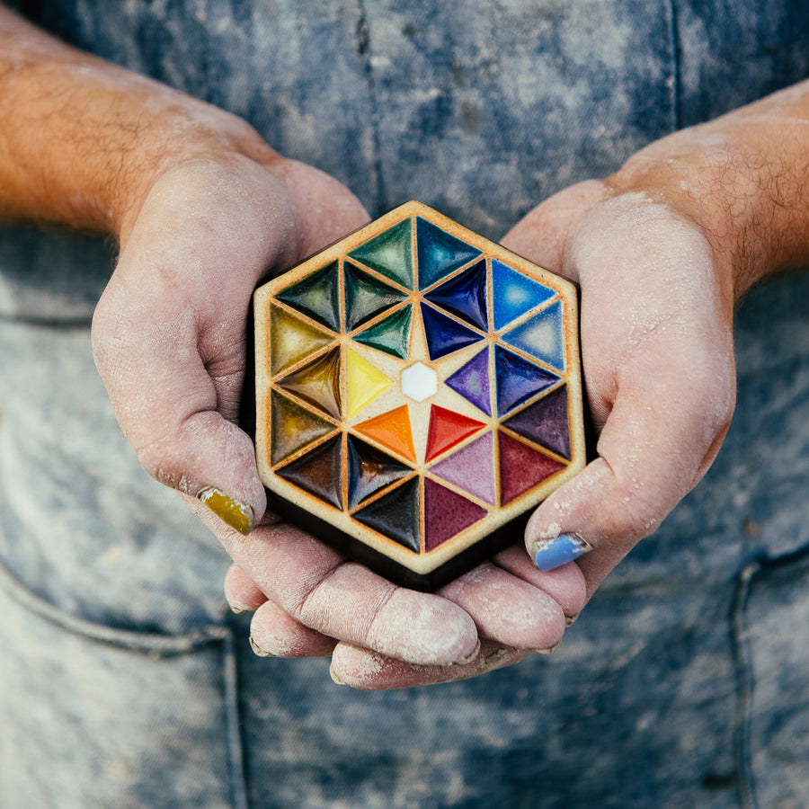 A Pewabic employee wearing a well-used denim apron holds the colorful Hex Paperweight in their dust-covered hands.