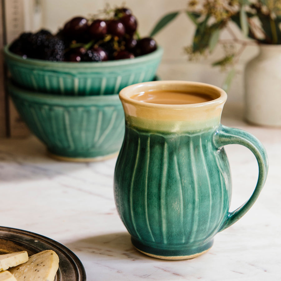 Classic Mug in Pewabic Green in the foreground filled to the brim with coffee and cream. In the background, there are two Small Pewabic Classic Bowls stacked on top of one another with blackberries and dark, red cherries.