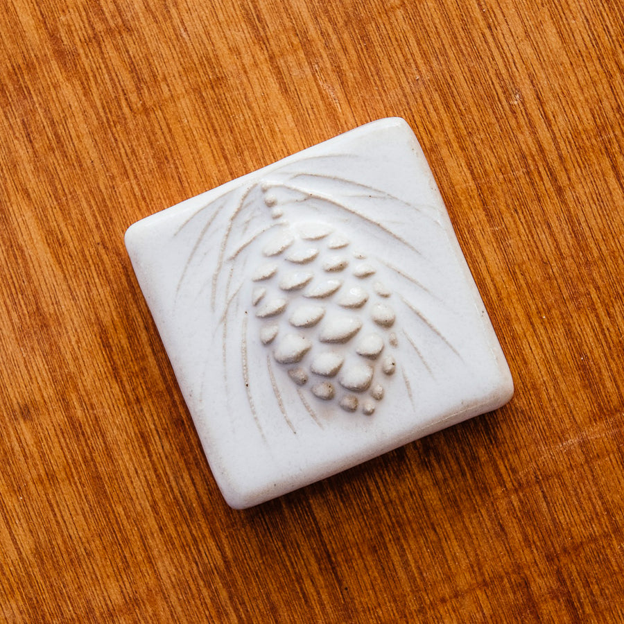 The ceramic Pinecone Tile has a high relief design with one large pinecone in the center and long spindly pine needles framing it.