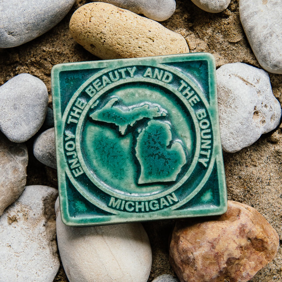 The 4x4 ceramic Michigan tile includes an embossed image of the two peninsulas of Michigan. Encircling these shapes are the words "Enjoy the Beauty and the Bounty, Michigan".