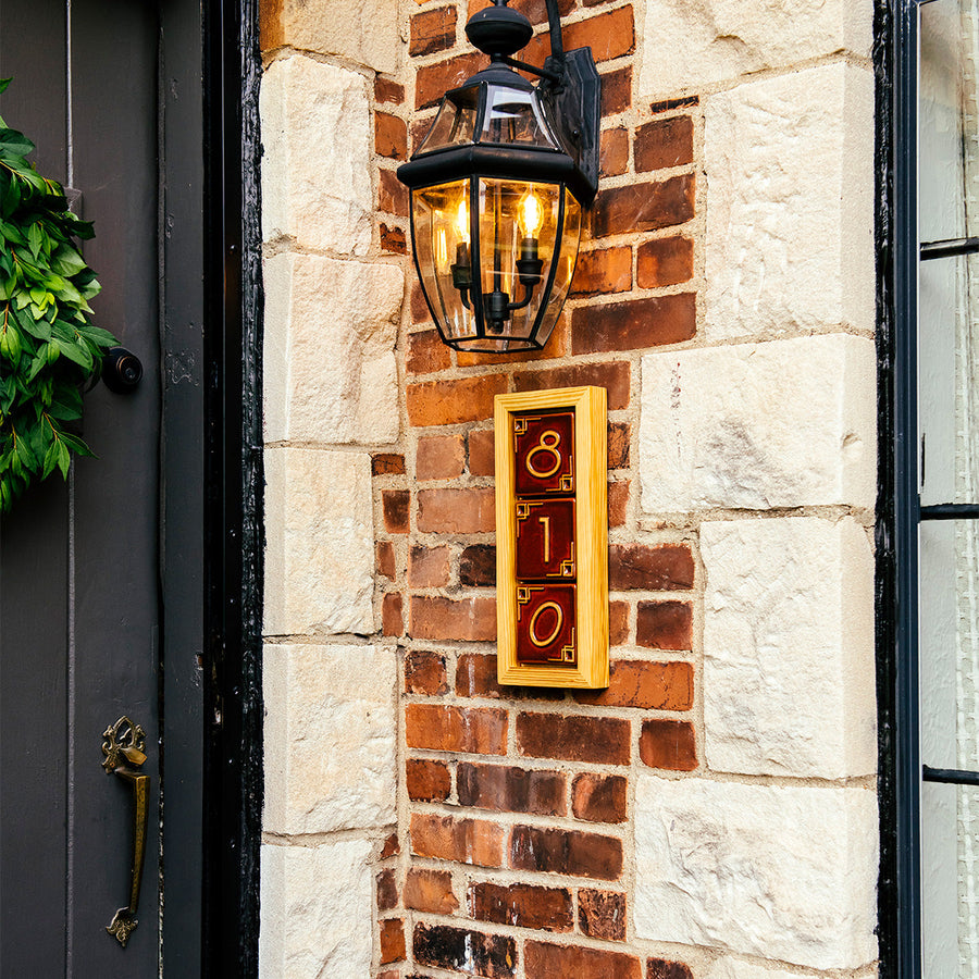 A three digit address frame is attached to a brick house below a porch light. The frame is made of a light blonde wood and holds brick red colored tiles.