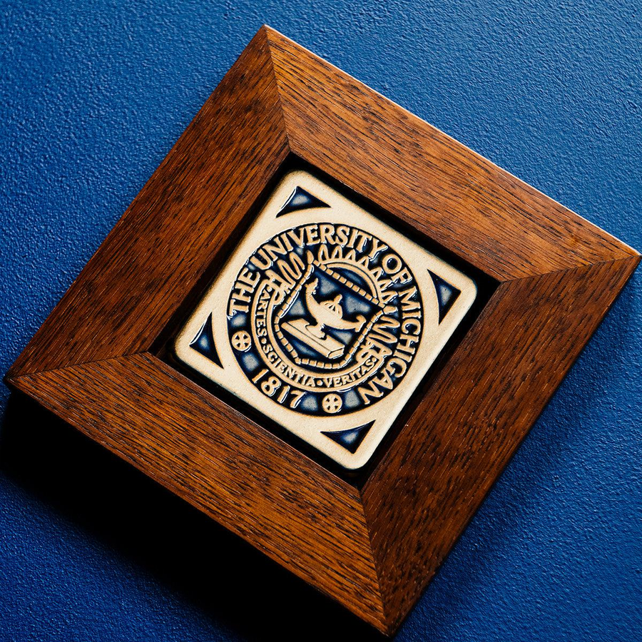 A 4x4 Midnight Scraped University of Michigan Seal tile in a rich wood frame rests on a royal blue surface