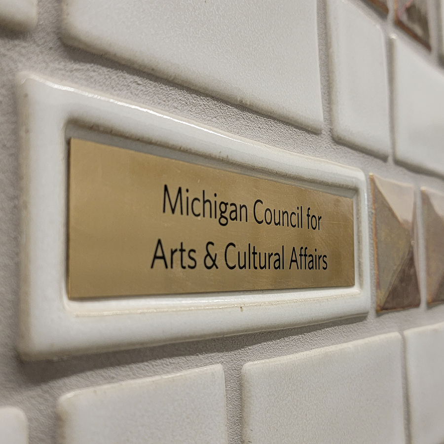 A detail of a donor wall features a gold plaque with the words "Michigan Council for Arts & Cultural Affairs" is surrounded by a white tile border.