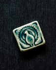 This small 2x2 tile features the image of a peacock facing the audience with its tail feathers open in a circular display. The tile is glazed in Lake Superior, a glossy greenish blue.