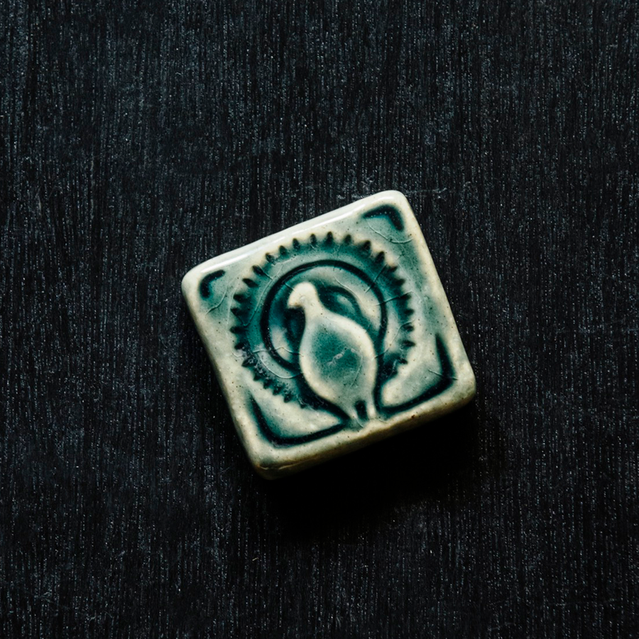 This small 2x2 tile features the image of a peacock facing the audience with its tail feathers open in a circular display. The tile is glazed in Lake Superior, a glossy greenish blue.