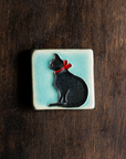This Cat Tile features a black cat wearing a bright red bow around its neck on a pale blue background.