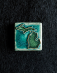 This hand painted tile has a matte turquoise background with matte earthy green peninsulas.