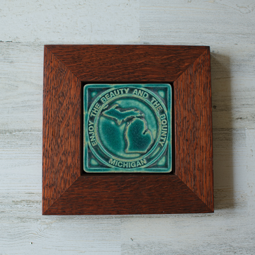 The 4x4 ceramic Michigan tile includes an embossed image of the two peninsulas of Michigan. Encircling these shapes are the words "Enjoy the Beauty and the Bounty, Michigan". The tile features the matte turquoise Pewabic Blue glaze which beautifully offsets the deep reddish brown of the oak wood frame.