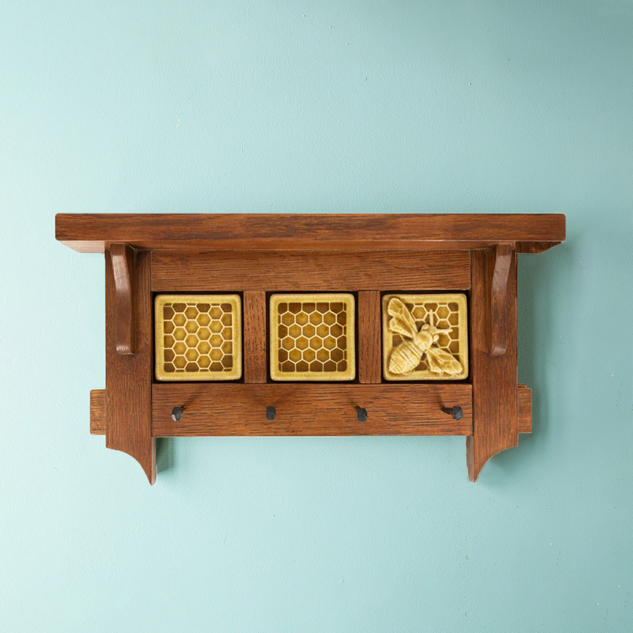 This triptych key rack features one Honey Bee Tile and two Honeycomb tiles. The Honeybee Tile features a large honeybee with stripes and detailed segmented wings sitting on a honeycomb patterned background. The Honeycomb tiles are a flat line-drawing of a honeycomb pattern with a simple smooth border around it. These three tiles are lined up and attached in a brown oak key-rack. The key-rack has a small shelf at the top and four black iron nails stick out of the bottom to hang your keys on.