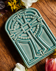 The Cross Tile has rounded edges at the top, creating a look similar to a stained glass window. The cross itself is ornate with a halo behind it. This tile features the glossy pale blue Glacier glaze.