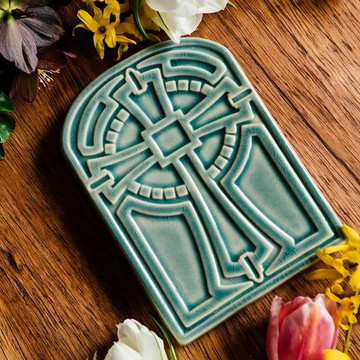 The Cross Tile has rounded edges at the top, creating a look similar to a stained glass window. The cross itself is ornate with a halo behind it. This tile features the glossy pale blue Glacier glaze.