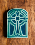 The Cross Tile has rounded edges at the top, creating a look similar to a stained glass window. The cross itself is ornate with a halo behind it. This tile features a two tone glaze with a pale blue cross and a deeper blue background.
