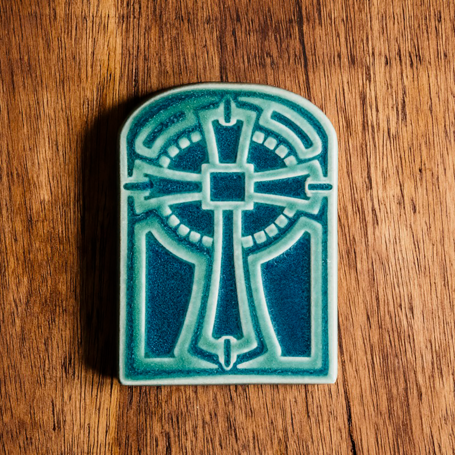 The Cross Tile has rounded edges at the top, creating a look similar to a stained glass window. The cross itself is ornate with a halo behind it. This tile features a two tone glaze with a pale blue cross and a deeper blue background.