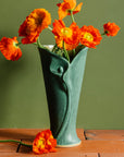 Bright orange poppies whimsically positioned in varying directions inside a Pewabic Calla Vase. The vase is featured in a matte, textured, green "Elm" glaze and rests upon terracotta roof tiles with a verdant green background.  
