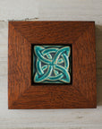 The Lover's Knot Tile features a central ring with four corner loops weaving around the ring from the corners to the center of the tile. This tile is in our matte turquoise Pewabic Blue glaze which beautifully offsets the deep reddish brown oak wood frame.