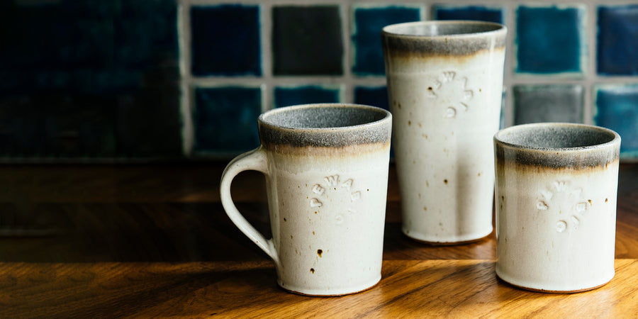 Arrangement of our Cafe Mug, Pint, and Rocks Cup all in a white, "Birch" glaze. They are set on a wooden table with an intricate grain. Varying blue glazed Pewabic Tile is in the background.