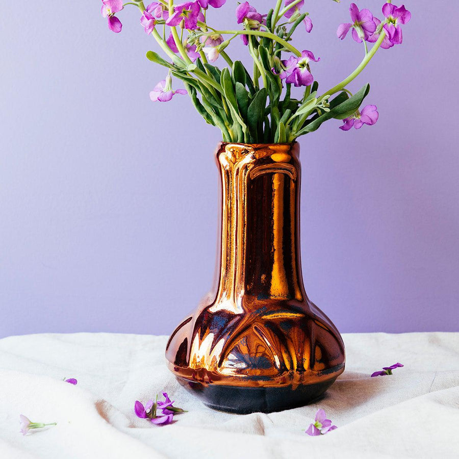 The Celtic Vase's base starts out wide and round at the bottom then becomes a straight pillar as it continues upward with a winding line design that has no beginning or end. The Copper iridescent glaze is a shiny, smooth metallic glaze with many variations. Some pieces end up with a blue-gray flashing of color that covers a portion of the surface.