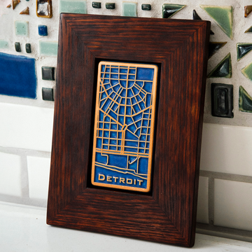 This rectangular Detroit Map Tile has a line drawing of the areal street view of downtown Detroit. The word "Detroit" is written at the bottom of the design. The raised areas of this tile - the word Detroit and the street lines - are scraped which means that it has an orange color while the background is a dark matte blue glaze. The frame is a deep reddish brown oak wood.