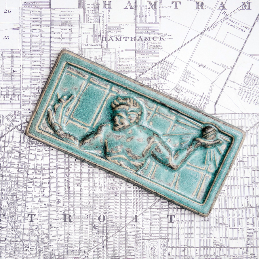 This high relief rectangular tile features a detail of the Spirit of Detroit sculpture found in downtown Detroit. The figure's arms are outstretched with a gilded sphere emanating rays in his left hand and a miniature family in his right. His head is bowed towards the family. The tile has a simple border. This tile features the mottled pale green Patina glaze. It has a similar look to the weathered green finish of copper.