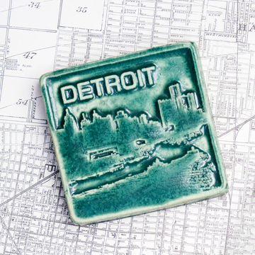 The Detroit Skyline tile features the current downtown skyline with the Detroit River in the foreground. A freighter floats by causing ripples on the otherwise flat surface of the water. The word "Detroit" is written above in the sky. This Detroit Skyline Tile features the matte blueish-green Pewabic Green glaze.
