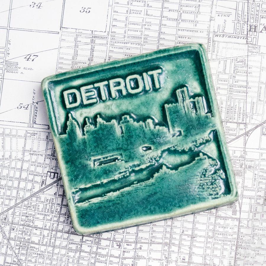 The Detroit Skyline tile features the current downtown skyline with the Detroit River in the foreground. A freighter floats by causing ripples on the otherwise flat surface of the water. The word "Detroit" is written above in the sky. This Detroit Skyline Tile features the matte blueish-green Pewabic Green glaze.