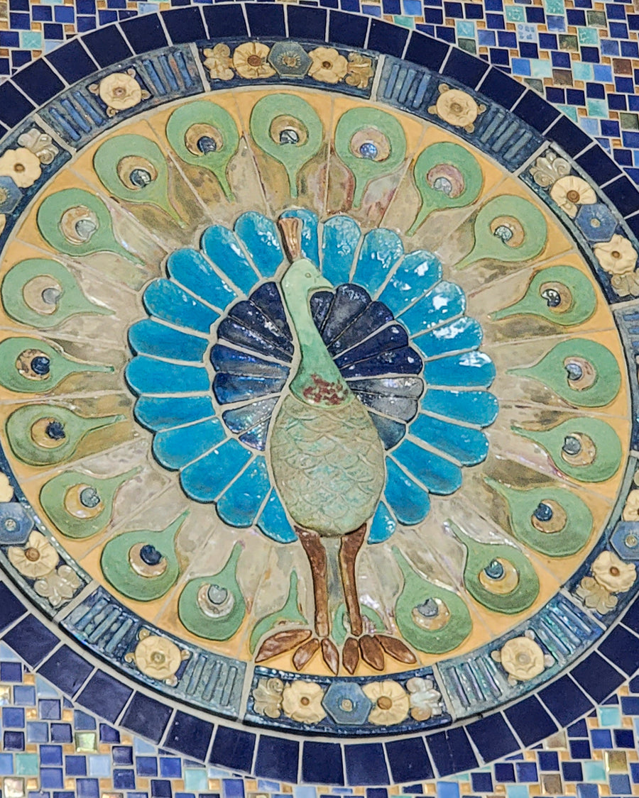 A detailed photo of the iconic peacock lunette fixed above the doorway to the Bird House at the Detroit Zoo. The peacock is set within a circular shape, with distinctive and delicate flower motif tiles bordering the design.