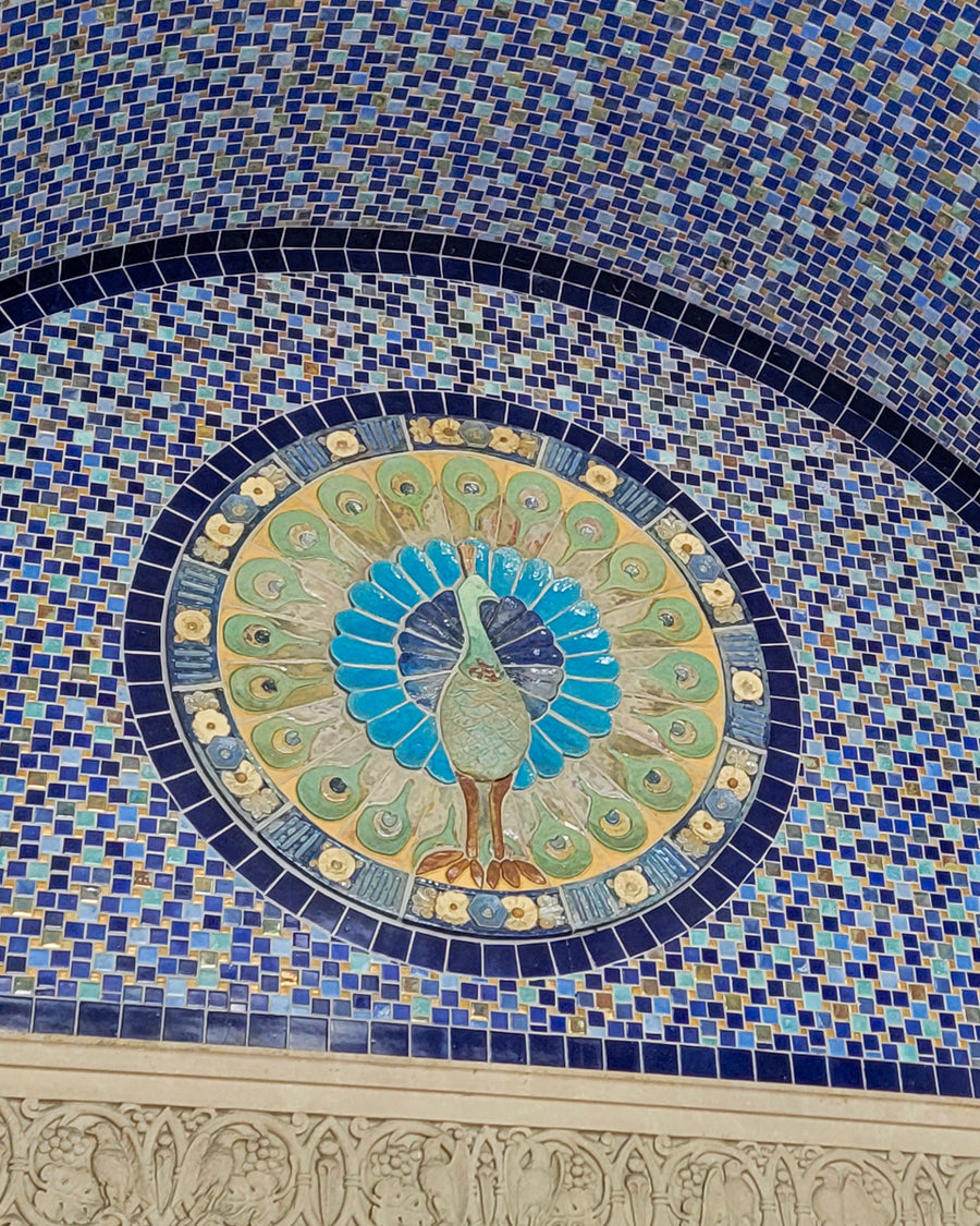 A detailed photo of the iconic peacock lunette fixed above the doorway to the Bird House at the Detroit Zoo. The peacock is set within a circular shape, with distinctive and delicate flower motif tiles bordering the design. This photo also includes details of the blue and iridescent tiles encompassing the intricate peacock feature.