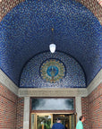 Two people are shown near the entrance of the Bird House at the Detroit Zoo in Royal Oak, Michigan. Pewabic tile in bright blues and golden iridescent line the domed entrance of the building. There is a prominent peacock motif lunette above the doorway completely made up of Pewabic tiles. 