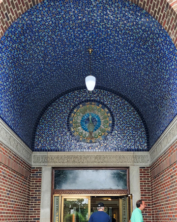 Two people are shown near the entrance of the Bird House at the Detroit Zoo in Royal Oak, Michigan. Pewabic tile in bright blues and golden iridescent line the domed entrance of the building. There is a prominent peacock motif lunette above the doorway completely made up of Pewabic tiles. 