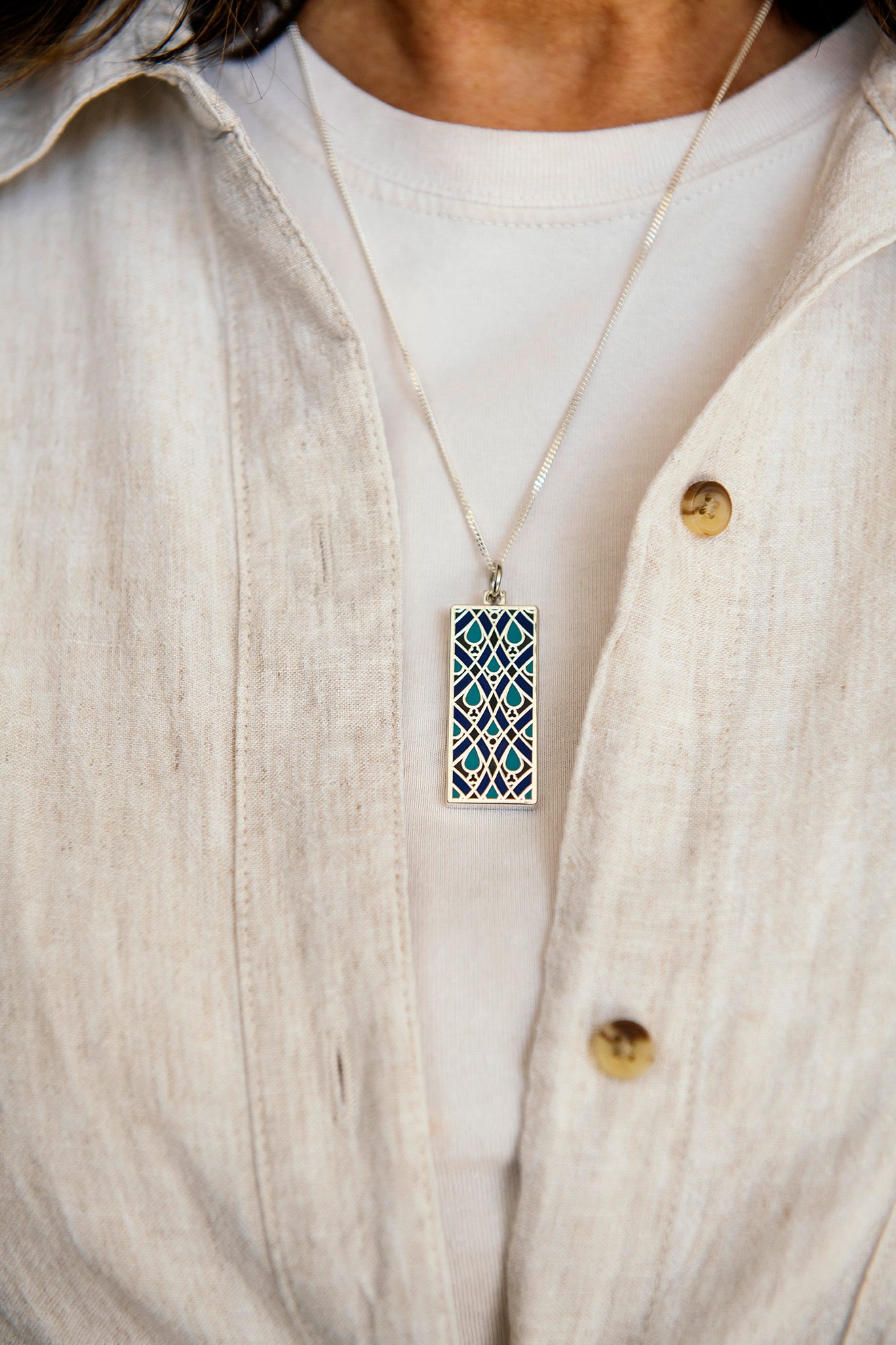 The Women's City Club necklace is a rectangular metal pendant with a geometric design that includes teardrop shapes and smoothly curved lines in various shades of blue. The necklace is worn by a person in a light-colored shirt with a linen button-up top.