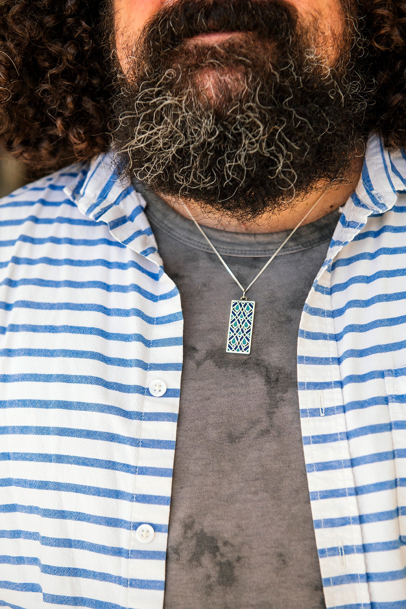 A man wearing a blue and white striped shirt paired with the Women's City Club necklace. The necklace features a rectangular metal pendant with a geometric design that includes teardrop shapes and smoothly curved lines in various shades of blue.