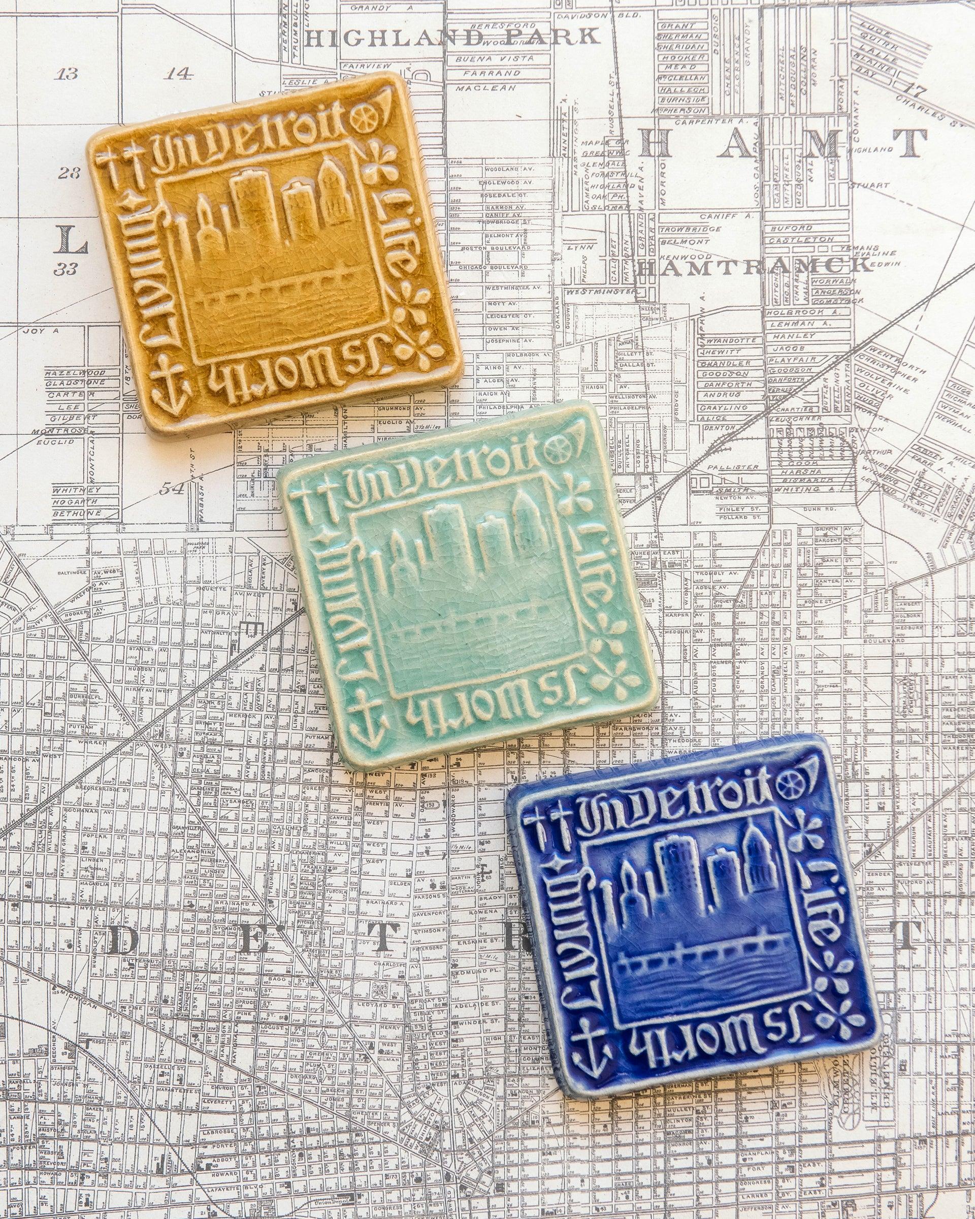 Three decorative Old Detroit tiles in glossy Honey, Celadon, and Lake Superior glazes on a map of Detroit background.  
