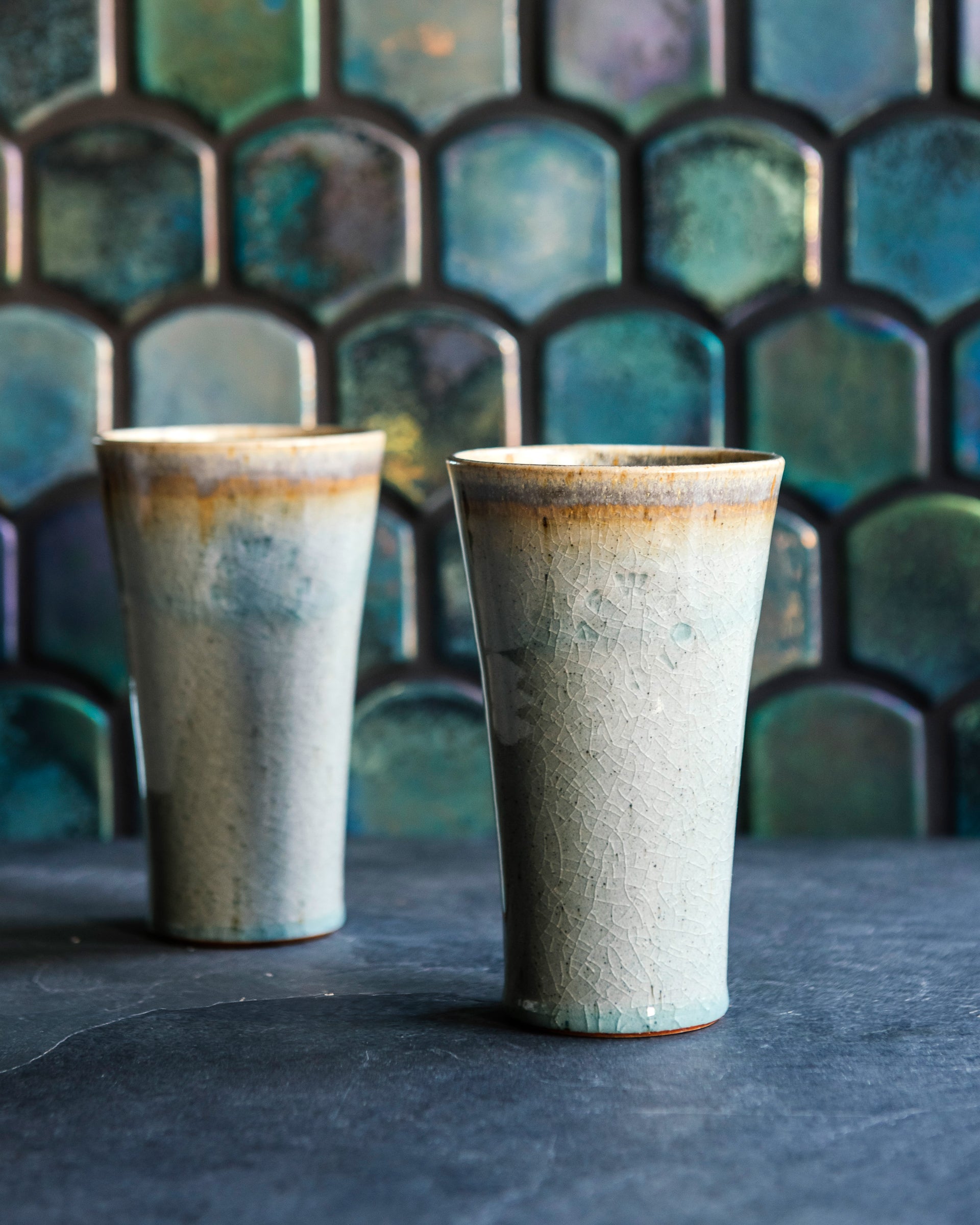 Two ceramic Pewabic Pints with a textured, glossy, ice-blue Frost glaze surface on a dark surface with a patterned Aurora Iridescent tile background.